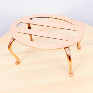 Puede incluir: Un salvamanteles de color cobre con una parte superior ovalada y tres patas curvas. La parte superior tiene un diseño ranurado. Este artículo decorativo está diseñado para proteger las superficies de los utensilios de cocina calientes.