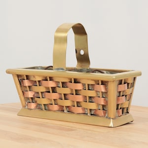 以下が含まれることがあります： 取っ手付きの真鍮と銅の織りバスケット。バスケットは長方形で、織り模様があります。