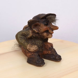 Puede incluir: Una figura de gnomo marrón y verde con barba gris y sombrero marrón. El gnomo está sentado con las piernas cruzadas y lleva botas marrones.