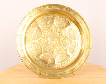 May include: A round, gold-colored metal tray with an intricate, embossed design. The tray has a raised rim and a decorative border.