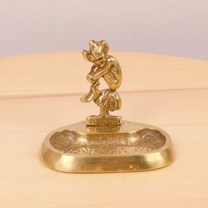 May include: A brass trinket dish with a small, detailed sculpture of a pixie sitting on a mushroom. The dish is inscribed with the word "Devon".