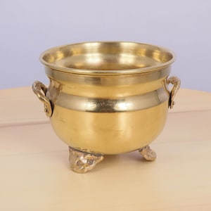 Puede incluir: Un pequeño macetero de latón de color dorado con dos asas y tres patas. El macetero está vacío y tiene una superficie lisa y brillante.