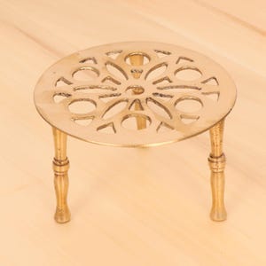Puede incluir: Un salvamanteles de metal dorado con una parte superior redonda decorativa y tres patas. La parte superior tiene un diseño recortado con un patrón floral.