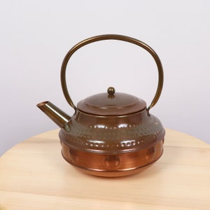 May include: A copper teapot with a hammered finish and a brass handle. The teapot has a round body and a lid with a knob. The teapot is sitting on a wooden surface.