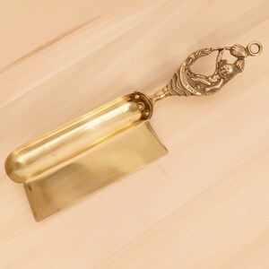 Può includere: Una paletta vintage in ottone con una lama rettangolare e un lungo manico curvo. Il manico presenta un rilievo dettagliato di una figura di folletto e il testo "Dartmoor Pixie". La paletta è probabilmente per uso in cucina o decorativo.