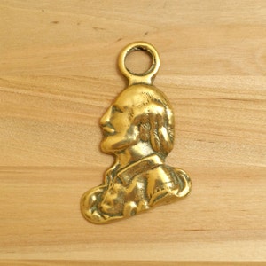 Puede incluir: Un tope de puerta de metal dorado con forma de cabeza de persona de perfil. El tope de puerta tiene un lazo en la parte superior para colgarlo.