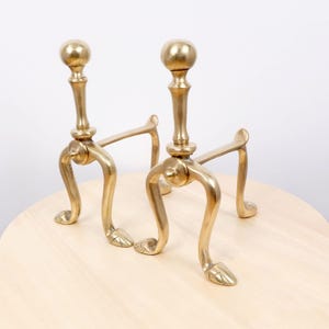 Puede incluir: Un par de morillos de latón a juego con un acabado metálico pulido. Cada accesorio de chimenea tiene un poste vertical rematado con un remate esférico, apoyado por tres patas curvas que terminan en pies detallados con forma de pezuña.