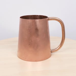 May include: A cylindrical copper mug with a smooth, metallic finish and a single curved handle attached to the side. The mug features a warm, reddish-brown hue and a consistent, polished surface texture throughout the entire exterior body.
