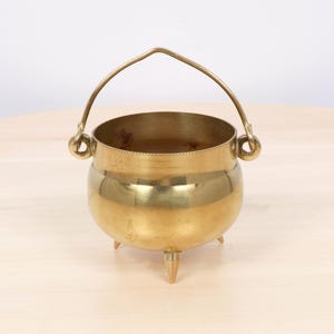 Puede incluir: Un caldero de latón pulido con cuerpo redondeado, tres patas puntiagudas y un asa móvil curvada sujeta a dos bucles laterales circulares. El interior del recipiente de metal muestra una pátina oscura, mientras que el exterior es liso.