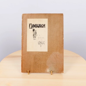 Pode incluir: Um livro antigo intitulado "Edinburgh" com um esboço de um tocador de gaita de foles na capa. O livro tem uma capa bege com um rótulo creme com o título e a ilustração. O livro é exibido em um suporte dourado.