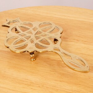 May include: A gold-toned metal trivet with a decorative, intricate design and a long handle. The trivet has three legs and is designed to protect surfaces from hot dishes.