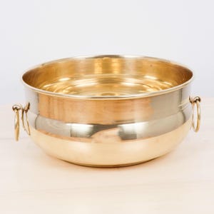 May include: A polished brass bowl with a smooth, rounded body and a wide, open top. The bowl features two circular brass ring handles attached to opposite sides, providing a functional grip. The metallic surface reflects light with a warm golden hue.