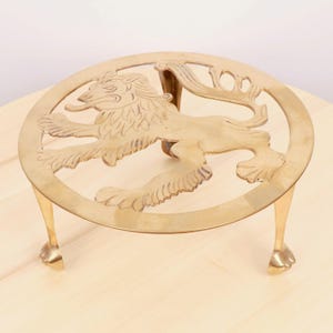Puede incluir: Un salvamanteles de metal dorado con una parte superior redonda y tres patas. La parte superior presenta un diseño recortado de un león.