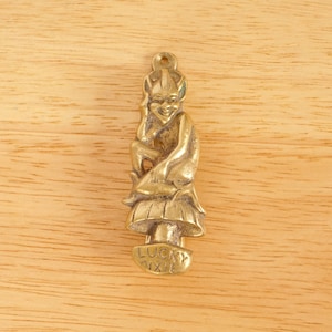 May include: A gold-colored brass doorstop shaped like a pixie sitting on a mushroom. The pixie is wearing a pointed hat and has a mischievous expression. The doorstop is inscribed with the words "Lucky Pixie".