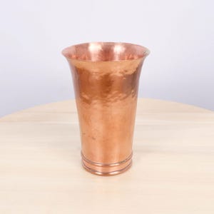 以下が含まれることがあります： フレアトップとハンマーテクスチャを備えた、背の高い銅色の花瓶。 花瓶は円筒形のボディと、ベース近くに2つの装飾リングがあります。 花瓶は明るい色の木製の表面に置かれています。