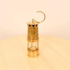 Könnte beinhalten: Eine Grubenlampe aus Messing mit einer Glasscheibe und einem Tragegriff. Die Lampe trägt ein Schild mit der Aufschrift "SAFETY LAMP OILTERY".