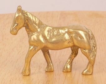 May include: A small, solid brass horse figurine. The horse is standing with its head held high and its legs slightly bent. The figurine is a light gold color and has a smooth, polished finish.