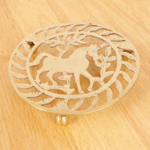 Op de afbeelding: Een gouden metalen onderzetter met een decoratief ontwerp van een paard in een cirkel. De onderzetter heeft drie poten.