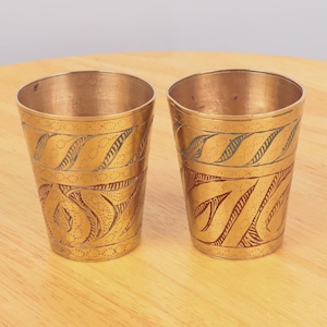 Puede incluir: Dos vasos de metal de color dorado con intrincados diseños grabados. Los vasos tienen un patrón rojo y verde.