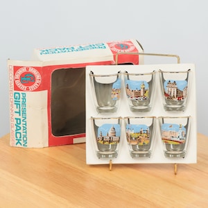 May include: A set of six clear glass shot glasses with gold rims. Each glass features a different image of a London landmark, including Big Ben, Buckingham Palace, and the Tower Bridge. The glasses are packaged in a white cardboard box with red and blue accents. The box reads "Presentation Gift Pack Decorated Drinking Glasses".