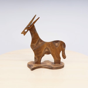 May include: A wooden donkey figurine with a clothespin head. The donkey is carved from wood with visible grain patterns and stands on a small wooden base. The figurine is a warm brown color and has a whimsical design.