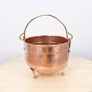 May include: A copper cauldron with a brass handle and three small brass feet. The cauldron has a rounded body and a shiny, reflective surface. The handle arches over the top, and the feet provide stability. The cauldron is likely used for cooking or decorative purposes.