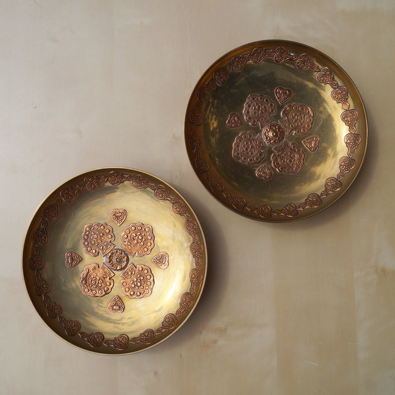 May include: Two decorative brass plates with intricate copper designs. The plates are round and have a raised rim. The designs are floral and symmetrical.