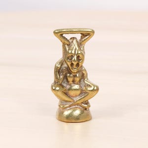 May include: A small, gold-toned brass figurine depicting a seated horned creature with pointed ears and a wide, open mouth. The figure is positioned in a cross-legged pose, resting on a rounded base, with a triangular opening at the top.