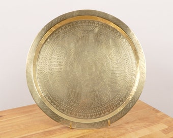 May include: A round, gold-colored metal serving tray with an intricate engraved design. The tray has a raised rim and a decorative border.