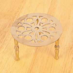 Op de afbeelding: Een gouden metalen trivet met een ronde, decoratieve bovenkant met een cirkelvormig, bloemmotief. De trivet heeft drie poten.