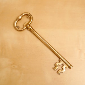 May include: A gold-colored skeleton key with a decorative head and a long, thin shaft. The key is lying on a light brown surface.