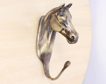May include: A bronze-colored horse head wall hook. The hook is shaped like a horse's head and neck, with a curved hook at the bottom. The horse head is mounted on a light-colored wooden surface.