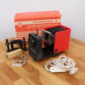 May include: A red and black slide projector with a white cord and a box labeled "спутник 2 guanpoekmon".