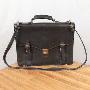 Puede incluir: Maletín de cuero negro con asa superior y bandolera desmontable. El maletín tiene un cierre de solapa con cerradura dorada y dos correas con hebilla. Las costuras blancas acentúan los bordes de la bolsa y la correa.