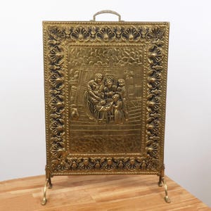 May include: A rectangular brass fireplace screen featuring an ornate border with repeating shell-like motifs. The central panel displays a relief scene of a group of figures gathered around a table. The screen includes a top handle and two legs.
