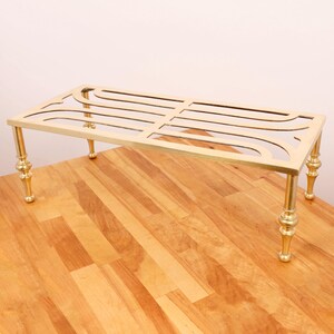 May include: A gold-toned metal stand with a decorative, curved design and four legs. The stand is made of brass and has a rectangular shape.