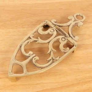 Op de afbeelding: Een gouden metalen trivet met een sierlijk ontwerp. De trivet is ovaalvormig met een decoratief handvat.