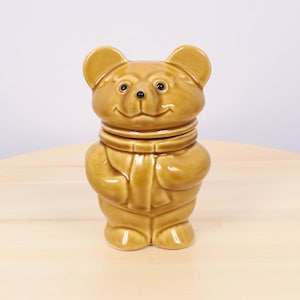 May include: A ceramic bear-shaped jar in a warm brown color. The bear has a smiling face, small black eyes, and a scarf-like detail around its neck. The jar is designed to stand upright and has a lid.