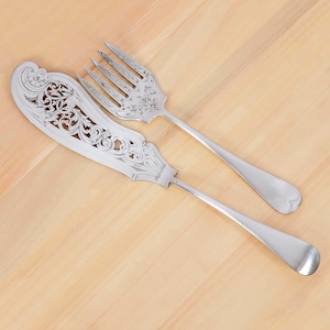 May include: A silver butter knife and fork set. The knife has an ornate, openwork design on the blade. The fork has four tines. Both pieces have long, slender handles and are set against a light wood background.