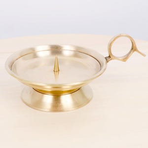 Puede incluir: Un candelero de latón con un asa circular y una espiga central. El candelero tiene una base redonda y un plato poco profundo para recoger la cera. El diseño es simple y elegante, adecuado para varios estilos de decoración.