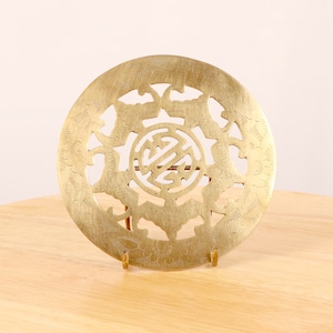 Puede incluir: Un posavasos redondo de latón con un diseño intrincado que presenta un símbolo central y motivos florales circundantes. El posavasos está sobre un soporte de madera.