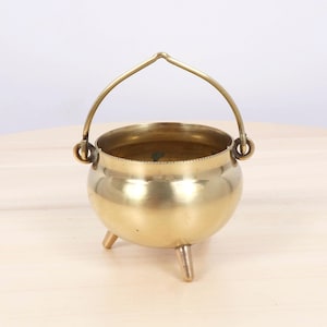May include: A polished brass cauldron featuring a rounded body, three short legs, and a curved handle attached to two circular side rings. The metal surface has a warm, golden tone with a smooth finish and a simple, open top design.