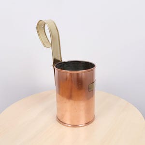 May include: A cylindrical copper vessel with a polished finish, featuring a tall, curved brass handle attached to the side. The container has a smooth, metallic surface and a small brass plate detail on the exterior wall, designed for hanging.