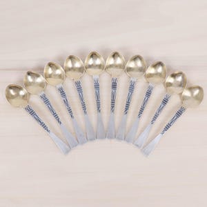 Puede incluir: Un conjunto de diez cucharas antiguas dispuestas en forma de abanico. Las cucharas tienen cuencos dorados y mangos plateados con un diseño geométrico decorativo. Se muestran sobre una superficie de madera clara.