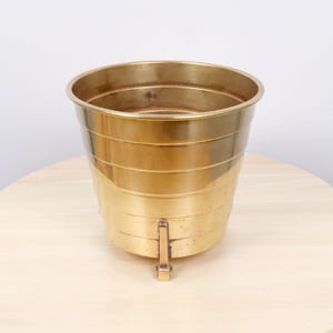 Può includere: Un portavaso in ottone lucido caratterizzato da un'ampia apertura superiore e da una serie di creste orizzontali a gradini lungo il corpo esterno. Il recipiente è sostenuto da tre piccoli piedini metallici verticali fissati alla base, oro.
