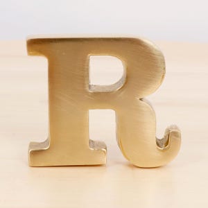 Könnte beinhalten: Ein goldfarbener Metallbuchstabe "R" mit gebürsteter Oberfläche. Der Großbuchstabe ist freistehend und steht auf einer hellen Holzoberfläche. Der Buchstabe ist etwa 10 cm hoch.