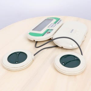 May include: A vintage Nintendo device, featuring a white and green handheld console connected by a gray cable to two white circular pads with dark green centers. The device is on a light wood surface.