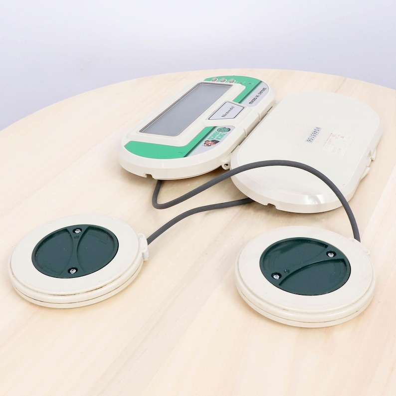 May include: A vintage Nintendo device, featuring a white and green handheld console connected by a gray cable to two white circular pads with dark green centers. The device is on a light wood surface.