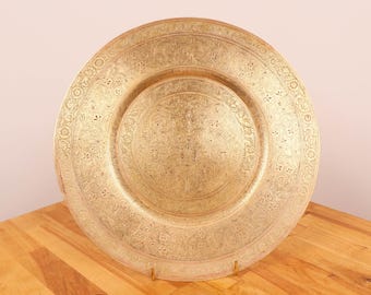 May include: A large, round, gold-colored metal charger plate with intricate floral designs etched into the surface. The plate is on a wooden surface.