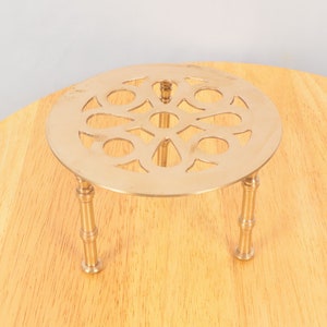 May include: A gold-toned metal trivet with a round, decorative top and three legs. The top has a cut-out design of a flower.